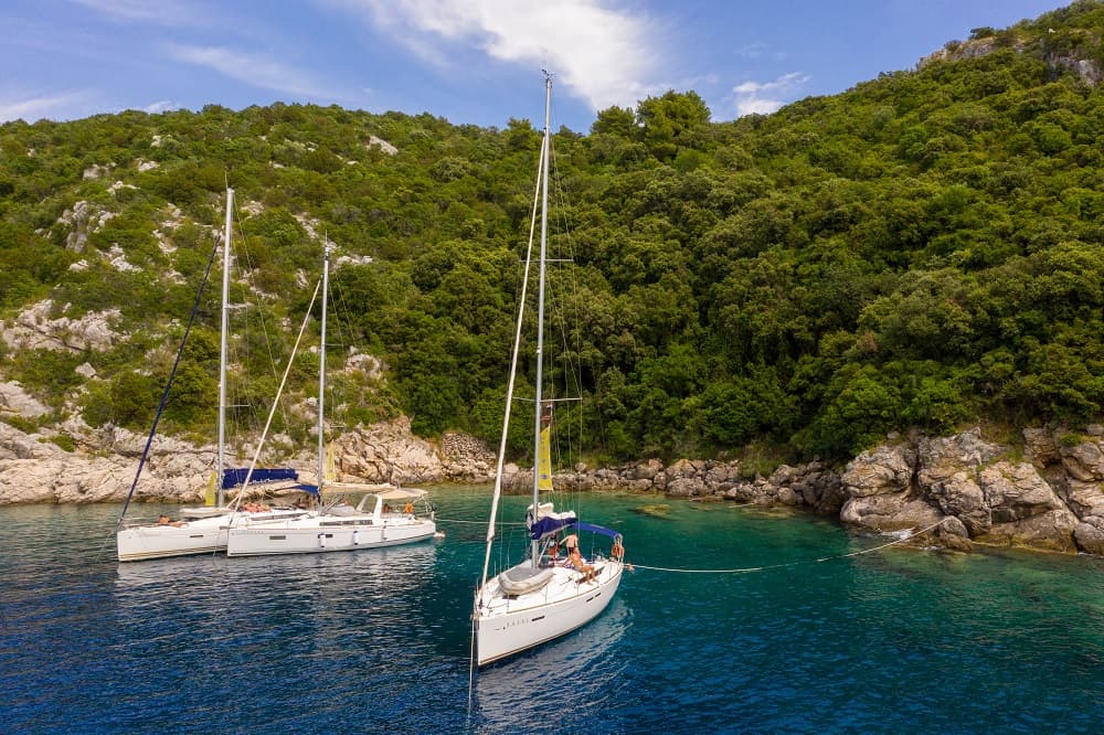Drei Segelboote ankerten während ihres Segeltörns unter einem strahlenden Himmel in einer Bucht mit klarem blauen Wasser neben einer üppigen, felsigen Küste.