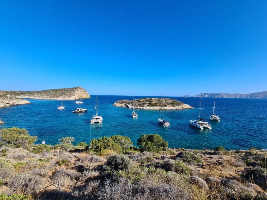 Ein malerischer Blick auf eine ruhige Bucht mit mehreren Segelbooten, die in kristallklarem blauem Wasser vor Anker liegen, umgeben von zerklüfteten Küsten unter einem strahlend blauen Himmel. Genießen Sie einen Segeltörn, bei dem Sie sich entspannen oder an Bord einer der ruhigen Segelyachten mitsegeln können.
