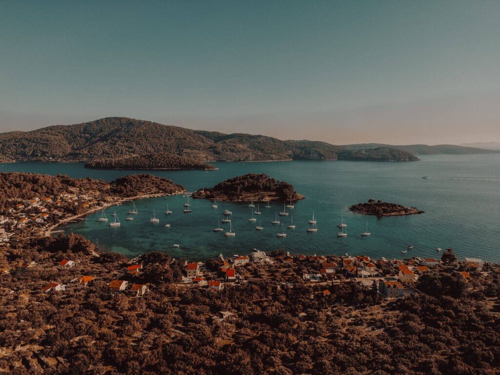 Luftaufnahme eines malerischen Küstendorfes mit rot gedeckten Gebäuden, umgeben von üppigem Grün, eingebettet neben einer klaren blauen Bucht, übersät mit Segelbooten auf einem Segelurlaub, mit bewaldeten