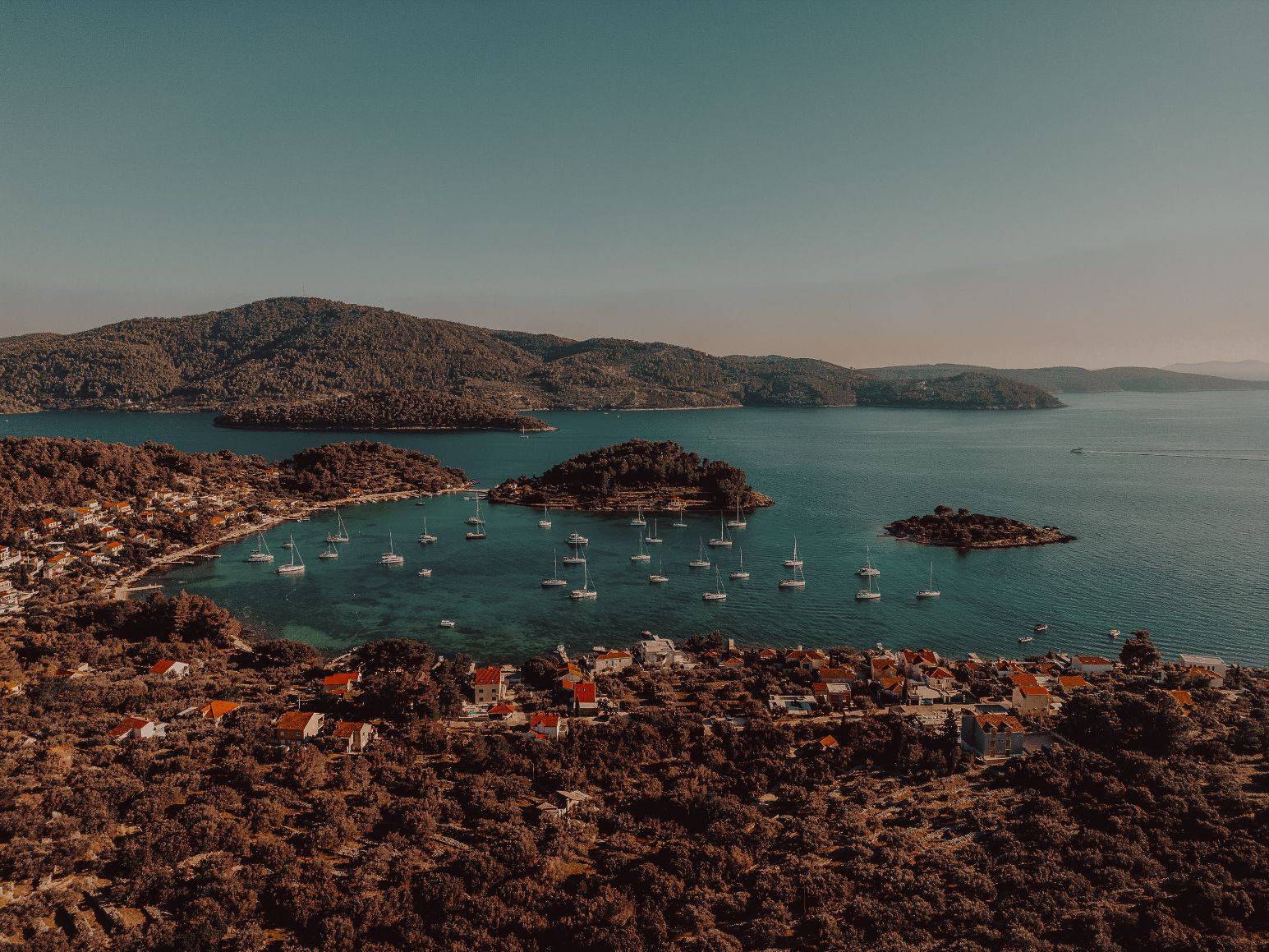 Luftaufnahme eines malerischen Küstendorfes mit rot gedeckten Gebäuden, umgeben von üppigem Grün, eingebettet neben einer klaren blauen Bucht, übersät mit Segelbooten auf einem Segelurlaub, mit bewaldeten