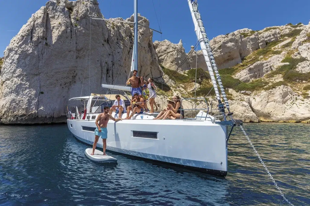 Eine Gruppe von Menschen genießt einen sonnigen Tag auf einem Segeltörn, der in der Nähe einer felsigen Küste vor Anker liegt. Einige sitzen auf dem Deck, während andere auf einem Paddleboard neben dem Boot stehen.