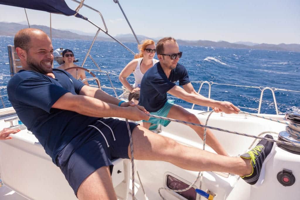 Drei Personen auf einer Segelyacht: ein lächelnder Mann am Steuer, ein anderer, der mühsam die Seile handhabt, und eine Frau, die zusieht. Alle tragen legere Segelkleidung an einem sonnigen Tag mit klarem blauen Himmel.