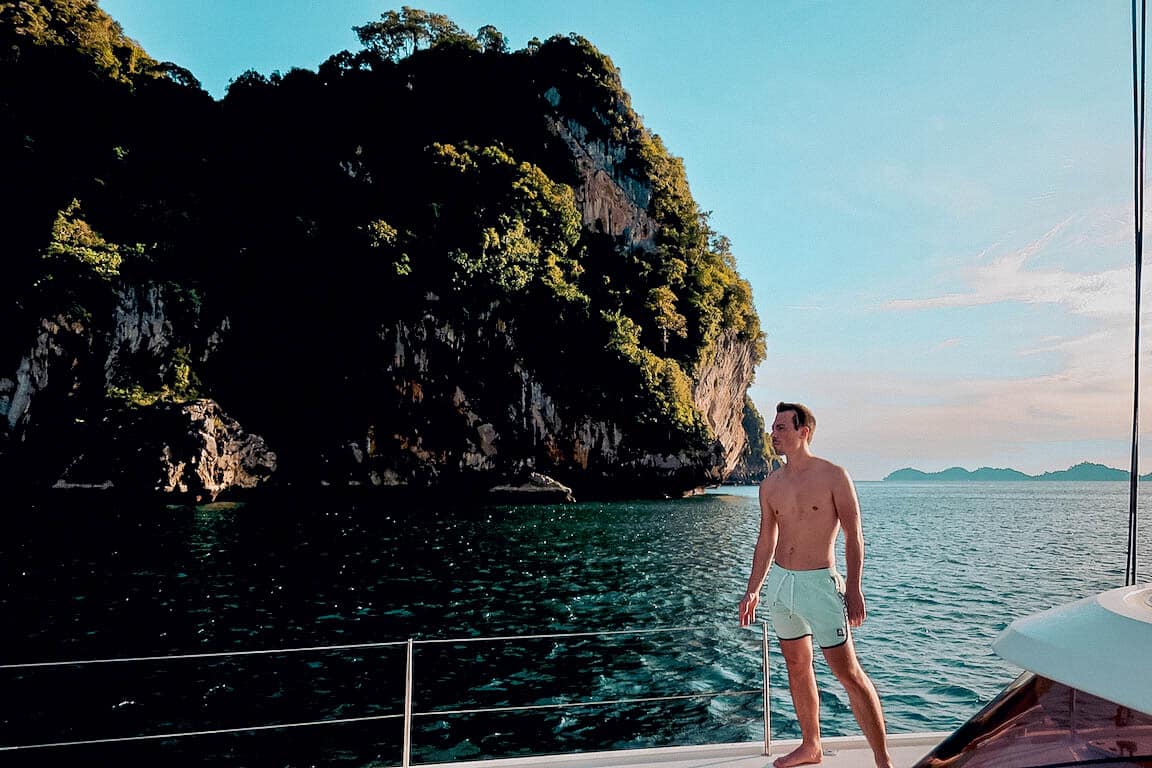 Ein Mann steht auf dem Deck einer Segelyacht und blickt auf eine große, üppige Insel unter einem klaren blauen Himmel. Der ruhige Ozean umgibt die Szene und schafft eine ruhige maritime Atmosphäre. Seine maßgeschneiderte Segelreise.