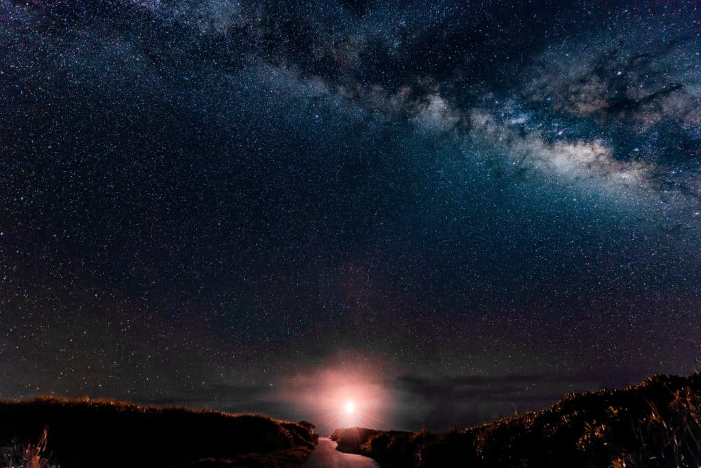 Ein weiter Nachthimmel voller Sterne und einer galaktischen Wolke über einer Landschaft, die vom hellen Licht eines Segeltörns am Horizont erhellt wird.