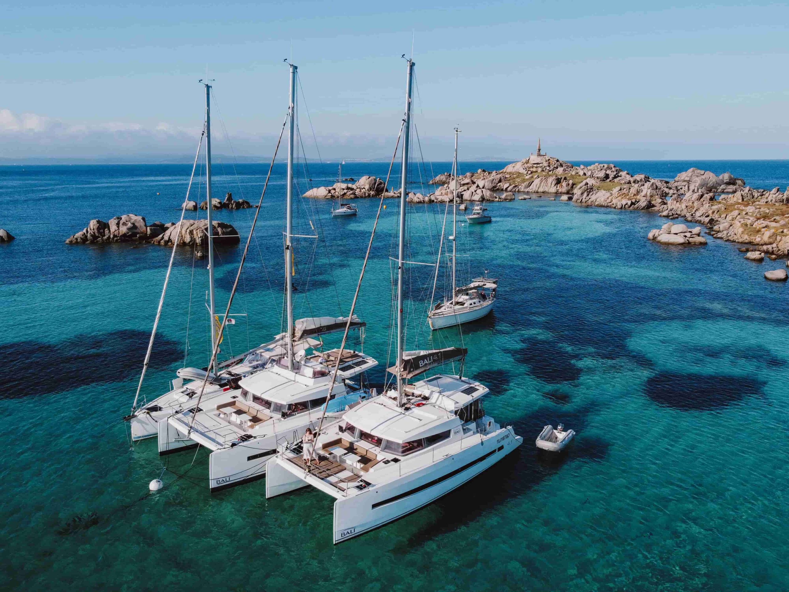Katamarane und Segelboote ankern in einer klaren türkisfarbenen Bucht mit felsigen Formationen im Hintergrund.