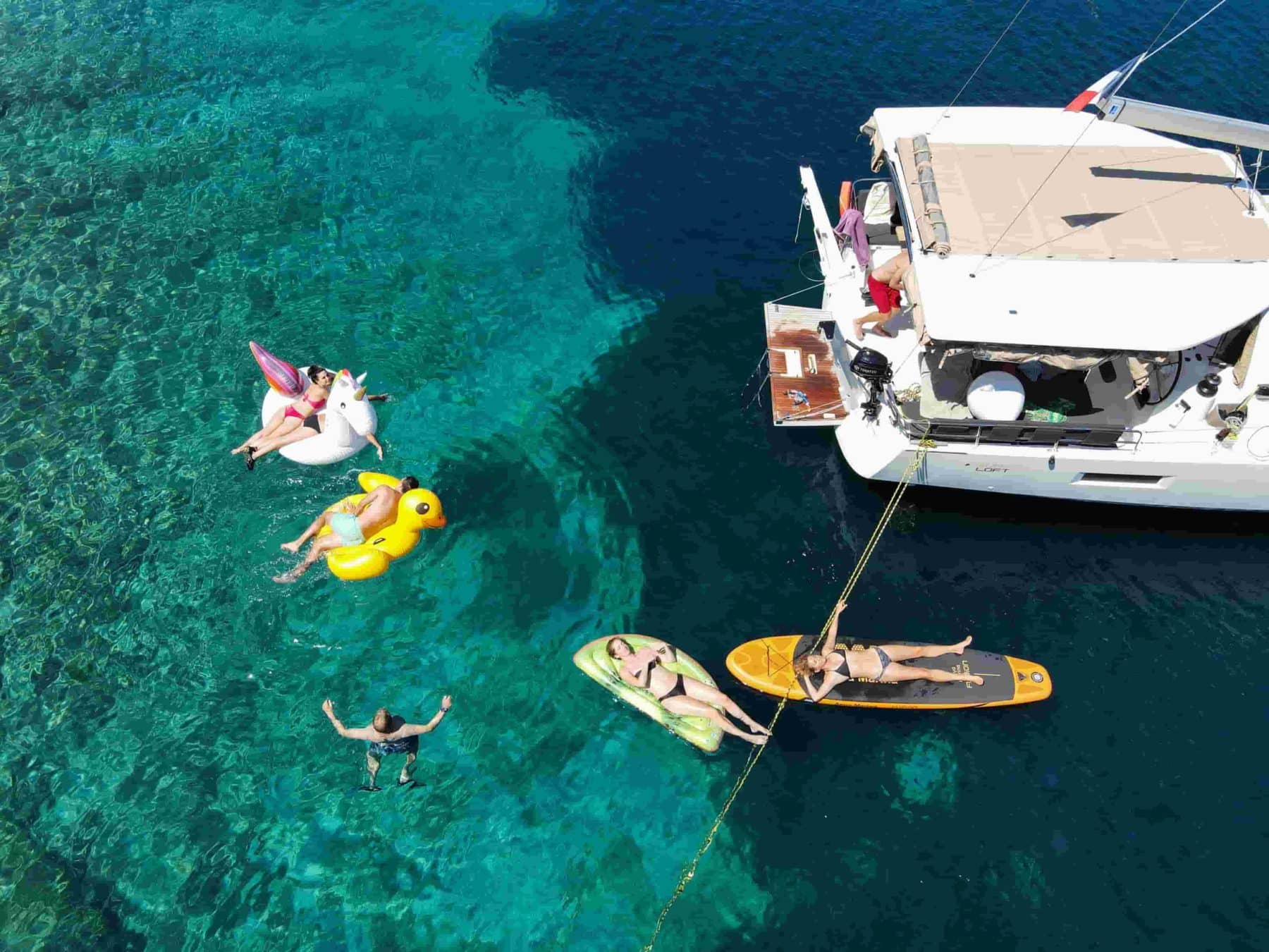 Luftaufnahme von Menschen, die Wasseraktivitäten rund um eine Segelyacht im kristallklaren Meerwasser genießen. Einige treiben auf aufblasbaren Spielzeugen, während andere schwimmen oder auf einem Paddleboard entspannen. 5 Aktivitäten Abenteuerurlaub