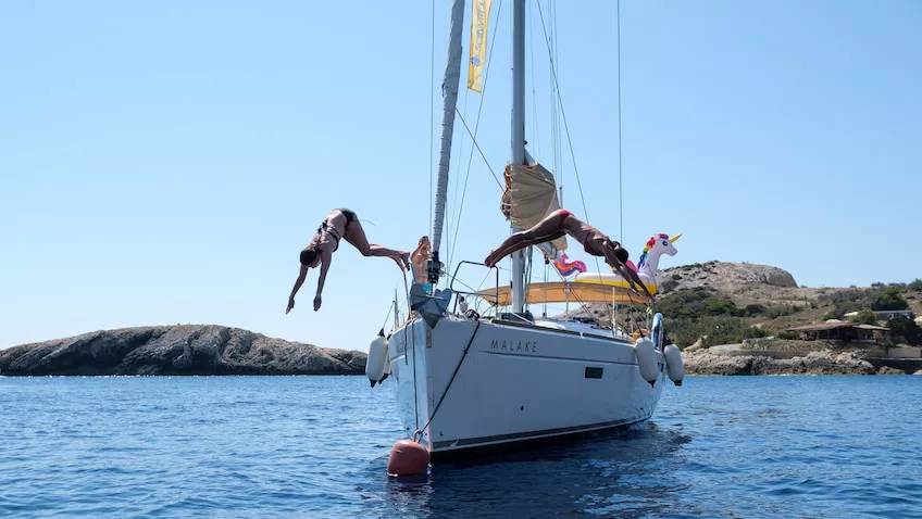 Unter einem klaren blauen Himmel springen zwei Menschen in der Nähe einer felsigen Küste von einer Segelyacht ins Meer. Abenteuerurlaub
