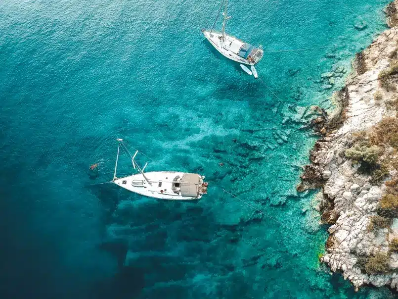 Luftaufnahme von zwei weißen Booten, die in der Nähe der felsigen Küste eines kristallklaren blauen Meeres vor Anker liegen, perfekt für einen Segeltörn.