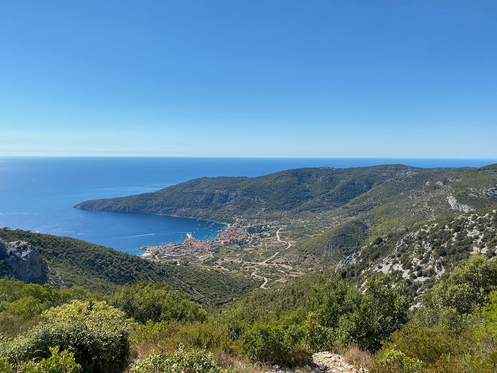Eine malerische Aussicht auf eine Küstenstadt, eingebettet zwischen üppig grünen Bergen und dem tiefblauen Meer mit seinen Segelyachten unter einem klaren Himmel.