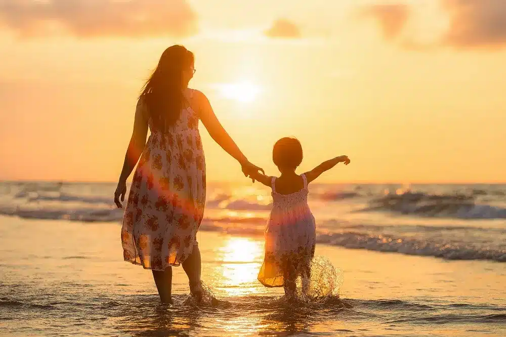 Eine Mutter und ihre Tochter gehen auf ihrer Segelreise Händchen haltend bei Sonnenuntergang durch das seichte Meerwasser und heben sich als Silhouette vor dem goldenen Himmel ab. Familien Badeurlaub tipps