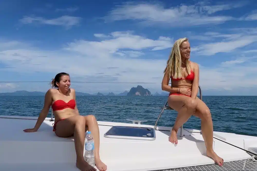 Zwei Frauen in roten Bikinis sitzen auf dem Deck eines Katamarans, lachen und genießen den sonnigen Tag mit einem malerischen Meer und felsigen Inseln im Hintergrund.