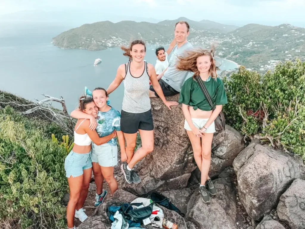 Eine fröhliche Gruppe von fünf Personen posiert während ihres Segeltörns auf einer felsigen Hügelkuppe mit Panoramablick auf eine üppige, grüne Küstenlandschaft und das ruhige Meer im Hintergrund. 5 Aktivitäten Abenteuerurlaub