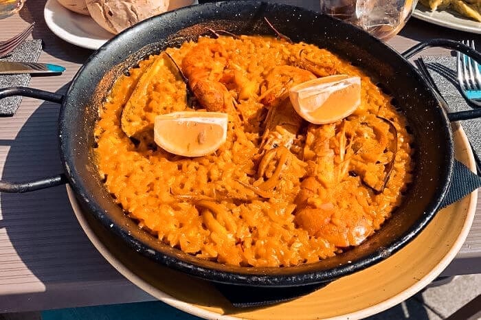 Eine schwarze Pfanne mit frisch gekochter Paella, darauf Reis, Garnelen und Zitronenscheiben. Das leuchtend gelb-orange Gericht steht auf einem Holztisch, im Hintergrund sieht man Besteck und Brot, was an ein köstliches Essen während eines Segelurlaubs auf einer luxuriösen Segelyacht erinnert.