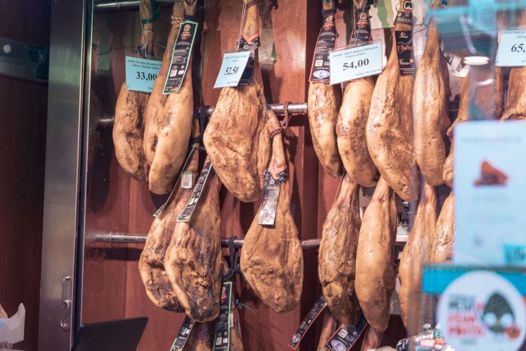 Eine Auswahl an luftgetrocknetem Schinken hängt während eines Segelurlaubs in einem Schaufenster, die Preisschilder sind in Euro angegeben.