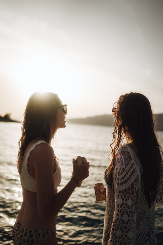 Zwei Frauen unterhalten sich während ihres Segelurlaubs bei Sonnenuntergang am Meer, eine hält ein Getränk in der Hand, beide werden von der Sonne im Gegenlicht beleuchtet, was ihre Silhouetten und den friedlichen Wasserhintergrund hervorhebt.