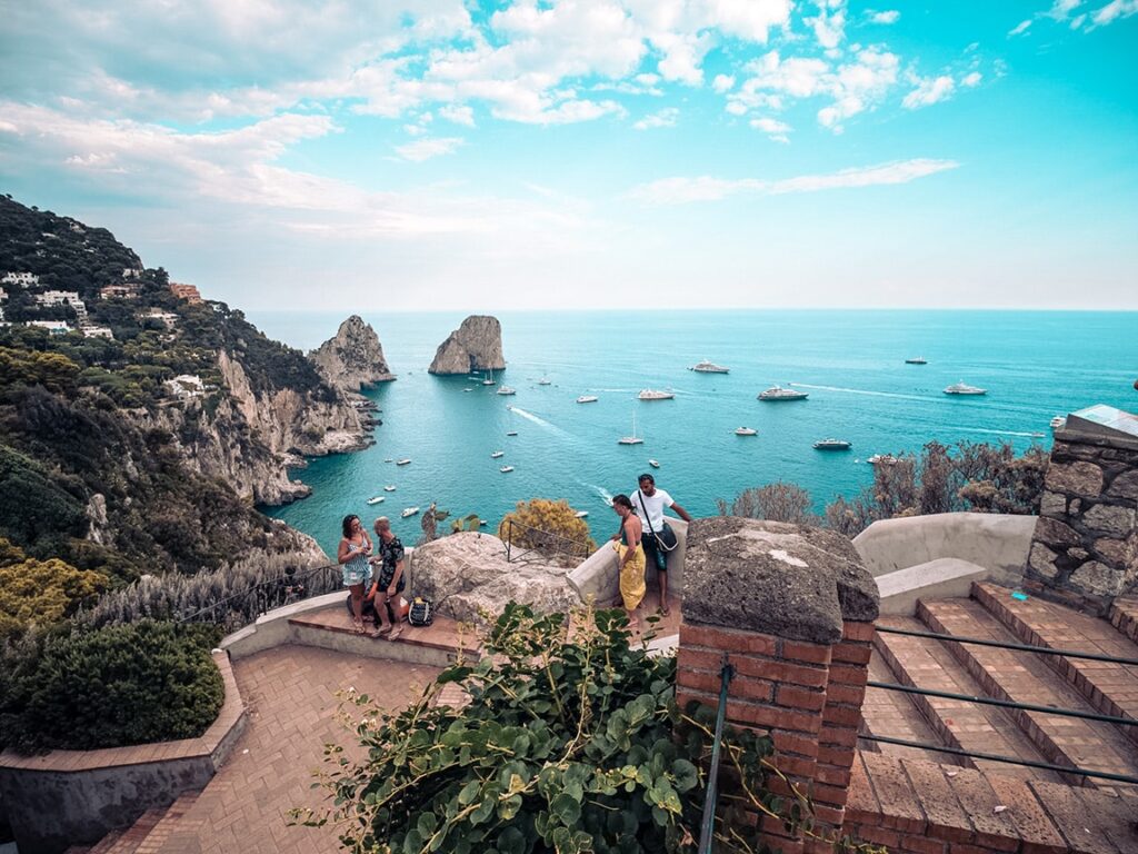 Touristen genießen eine malerische Aussicht von einem Aussichtspunkt an der Küste, mit üppigem Grün, blauem Meer, übersät mit Katamaranen und Felsvorsprüngen im Hintergrund unter einem teilweise bewölkten Himmel. Das Bild fängt eine lebendige und ruhige Küstenlandschaft ein, perfekt für einen Segelurlaub. besten Destinationen spontaner Kurzurlaub