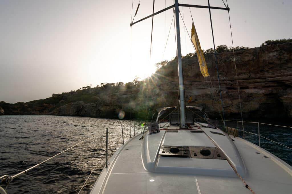 Der Bug eines Segelboots liegt in der Nähe einer felsigen Küste unter einem sonnigen Himmel. Das Sonnenlicht erzeugt im Bild einen Lichtreflexionseffekt.