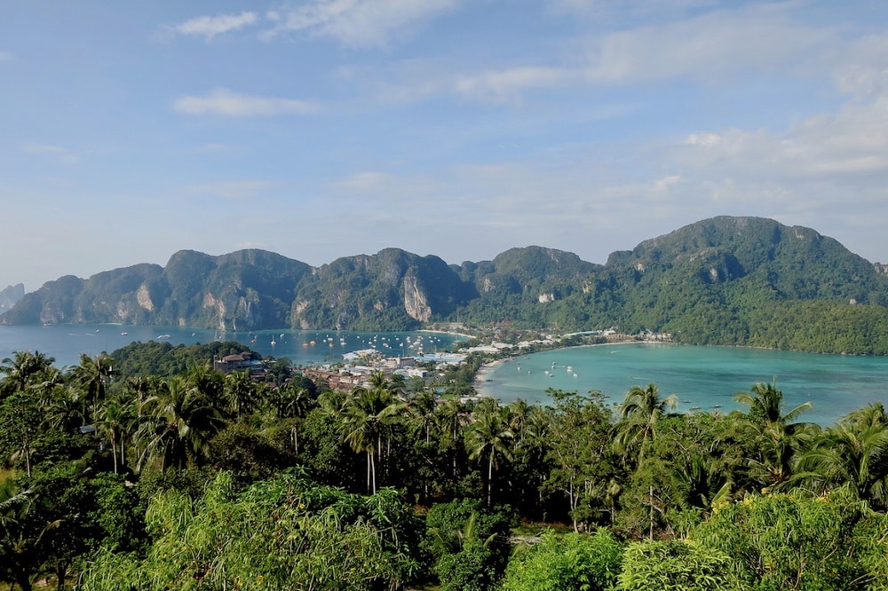 Ein Panoramablick auf eine tropische Küstenstadt mit üppig grünen Hügeln, klarem blauen Wasser und Segelyachten in Küstennähe unter einem strahlend blauen Himmel. Thailand Segeltörn als Geheimtipp