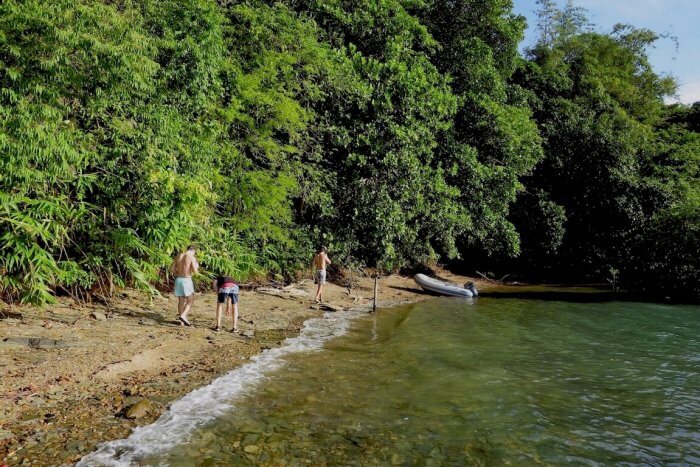 Drei Menschen gehen an einem Kieselstrand neben einem üppigen Wald entlang. Eine kleine Segelyacht liegt auf dem Sand nahe der Wasserkante. Thailand Segeltörn als Geheimtipp