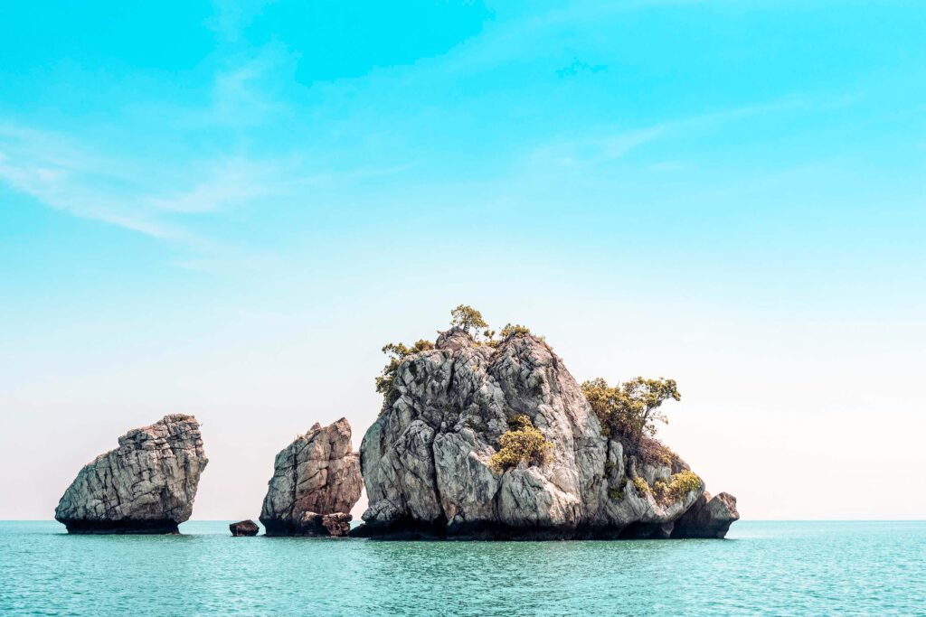 Eine ruhige Meeresszene mit drei großen Felseninseln, die in blaugrünem Wasser verstreut sind, perfekt für einen Segelurlaub. Die Felsen sind teilweise mit Sträuchern und kleinen Bäumen bewachsen. Der Himmel ist klar und strahlend blau mit einem sanften Farbverlauf zum Horizont hin und schafft so eine ideale Kulisse für Ihr Segelyacht-Abenteuer. in Thailand einsame ruhige entspannte bucht.