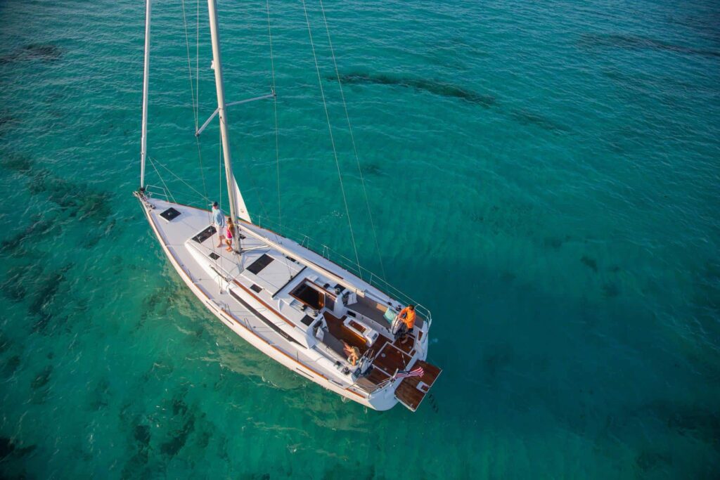 Luftaufnahme eines Segelboots mit weißen Segeln, das über klares, türkisfarbenes Wasser gleitet. An Bord der Segelyacht befinden sich zwei Personen, die den sonnigen Tag genießen. Das ruhige Meer darunter gibt dank der Klarheit des Wassers den Blick auf den Meeresboden frei und macht diese Segelreise zu einem perfekten Ausflug. Der perfekte Segelurlaub an Bord.