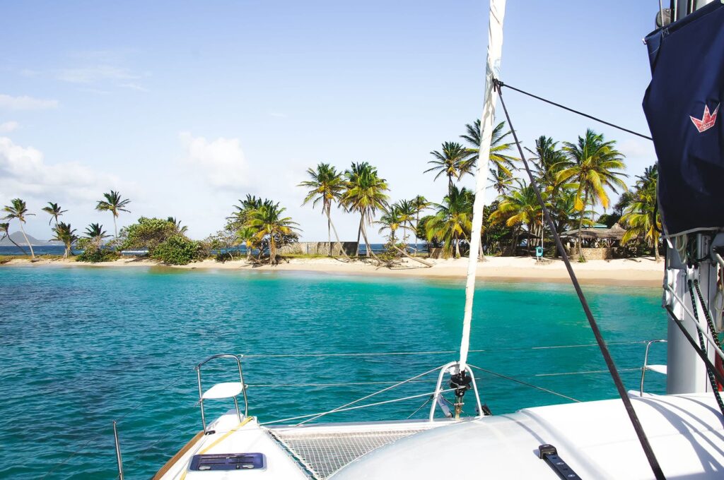 Eine Segelyacht liegt an einem tropischen Strand mit klarem, türkisfarbenem Wasser. Die Küste ist mit Palmen gesäumt, und im Hintergrund sind ein paar Hütten inmitten üppigen Grüns vor einem klaren, blauen Himmel zu sehen – ein Segelurlaub wie aus dem Bilderbuch.