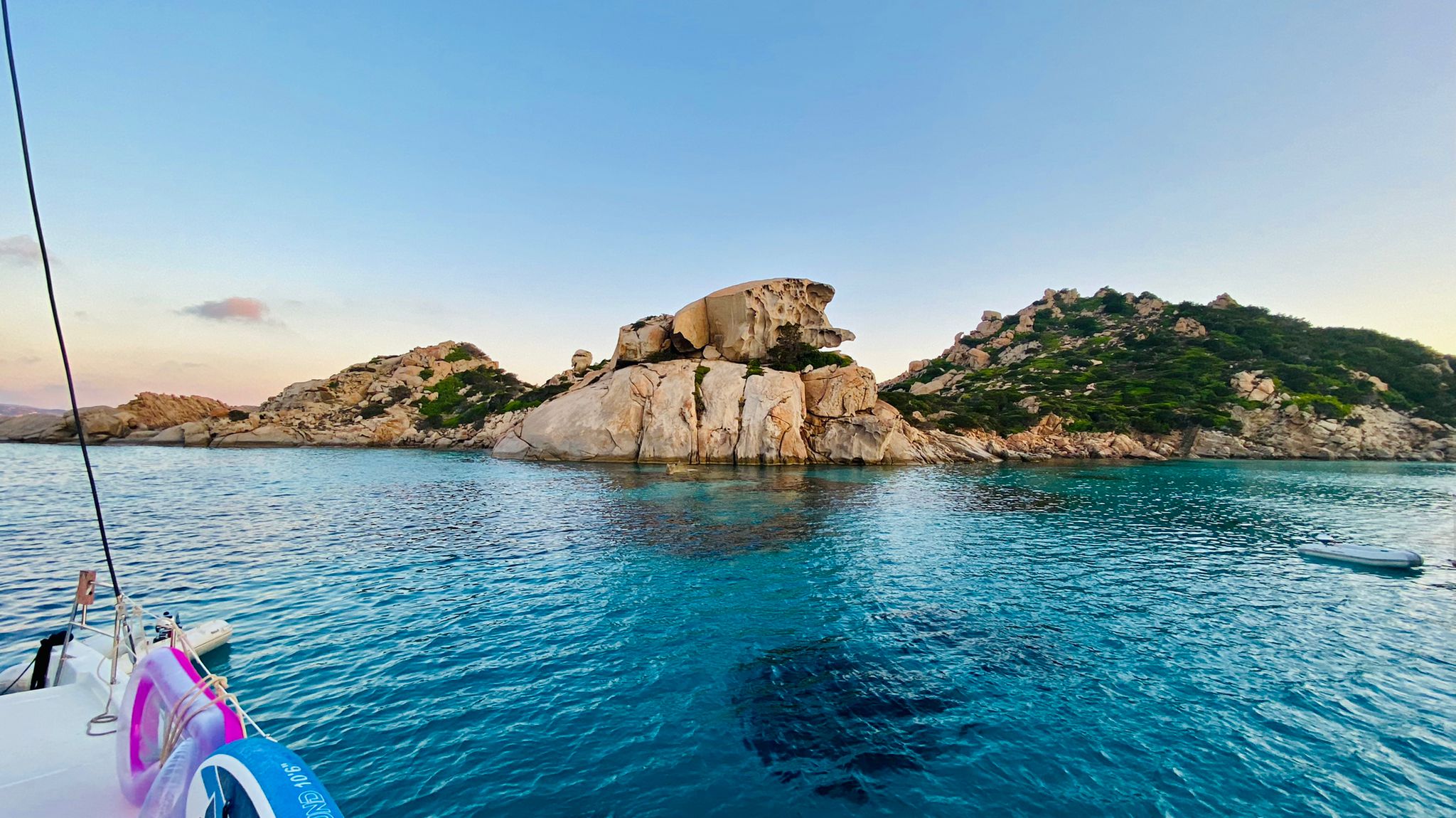 Es offenbart sich eine malerische Szene einer felsigen Insel, umgeben von klarem, türkisfarbenem Wasser. Im Vordergrund erinnert ein Bootsdeck mit einem rosa-weißen Rettungsring an einen abenteuerlichen Segeltörn. Der Himmel darüber ist klar mit einem Hauch von Sonnenuntergangsfarben, perfekt zum Mitsegeln an diesem ruhigen Abend.