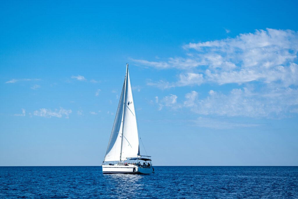 Ein weißes Segelboot mit voll ausgefahrenen Segeln segelt auf einem ruhigen blauen Meer unter einem klaren blauen Himmel mit ein paar vereinzelten Wolken. Im Hintergrund ist der Horizont sichtbar, der den perfekten Segelurlaub verkörpert und eine heitere und friedliche maritime Szene präsentiert.