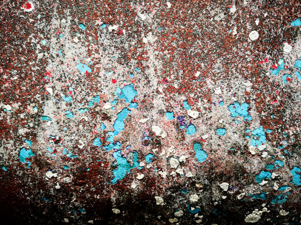 Eine Nahaufnahme einer strukturierten Oberfläche mit rostbraunem Hintergrund, die an ein abstraktes Kunstwerk erinnert, das vom Mistralwind geformt wurde. Es ist mit verschiedenen Farben gesprenkelt, darunter weiße Flecken, leuchtendes Blau und ein Hauch von Rot und Grün. Die Oberfläche wirkt abgenutzt und verwittert.