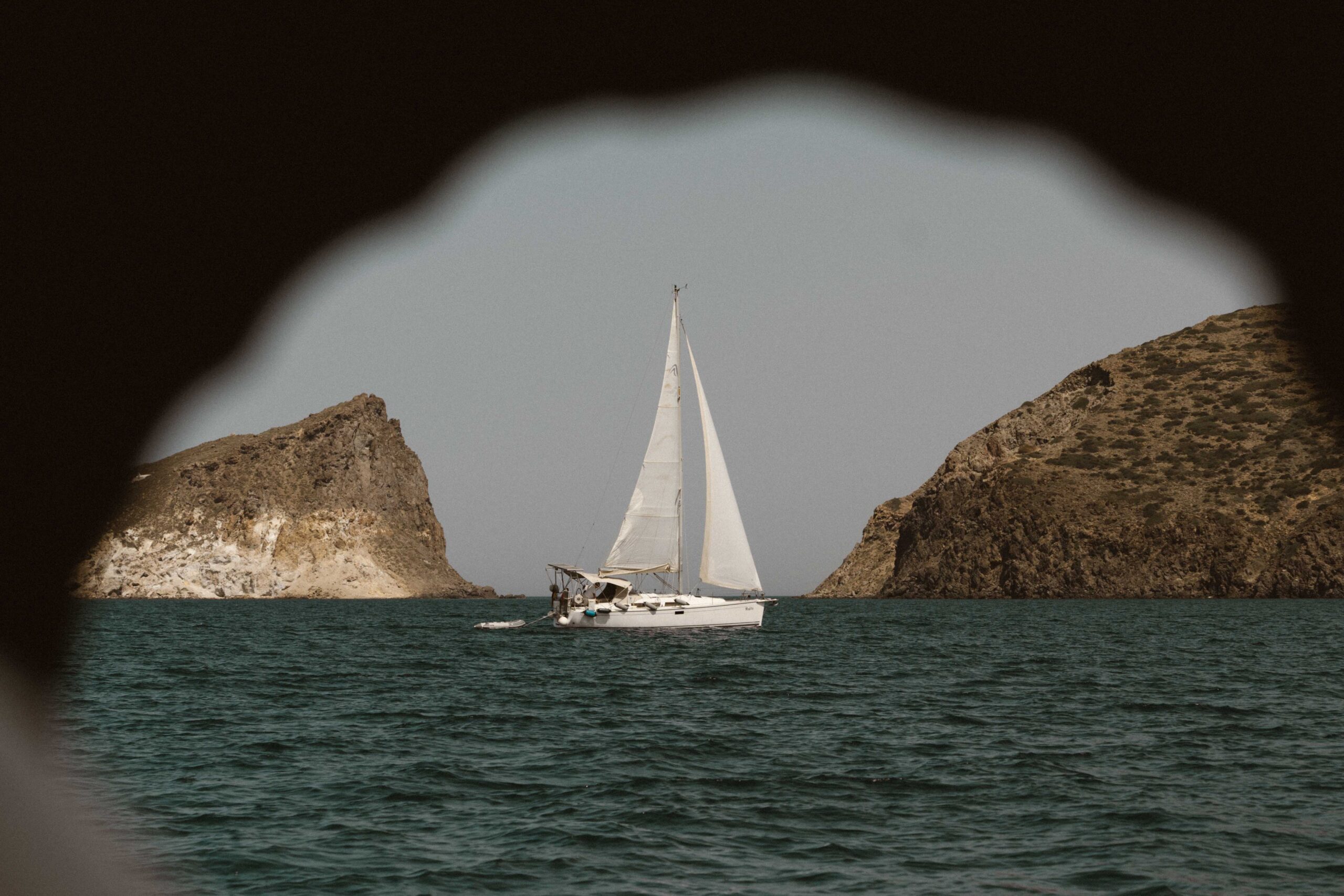 Ein weißes Segelboot gleitet auf einem ruhigen Meer, eingerahmt von felsigen Klippen auf beiden Seiten. Das Bild, das an ein Segelabenteuer erinnert, wird durch eine schlüssellochartige Öffnung betrachtet, mit dem Segelboot in der Mitte. Der Himmel ist klar und das Wasser ist tief blaugrün. Beste Wassersportart Badeurlaub und Segelurlaub.