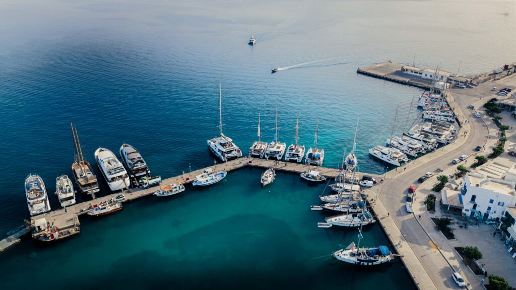 Luftaufnahme eines Küstenhafens auf den Kykladen. Man sieht zahlreiche Boote und Yachten, die an den Piers im klaren blauen Wasser angedockt sind, und in der Ferne sind ein paar kleine Boote zu sehen. Die Küste ist von Gebäuden und einer Straße mit geparkten Autos geprägt. Ein kleines Boot verlässt den Hafen.