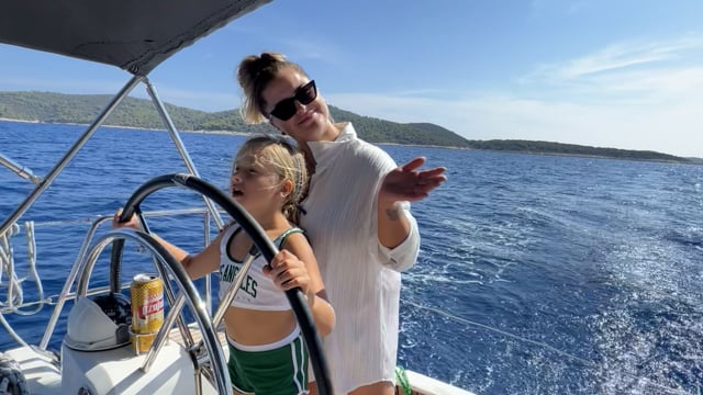 Ein Erwachsener und ein lächelndes Kind in grün-weißer Sportkleidung sind auf einem Segelboot. Das Kind hält das Steuerrad, während der Erwachsene spielerisch gestikuliert. Sie sind umgeben von blauem Meerwasser mit grünen Hügeln im Hintergrund. Es ist ein sonniger Tag.