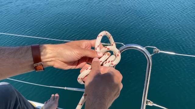 Person bindet mit einem weißen Seil mit roten und schwarzen Flecken einen Knoten an einer Bootsreling. Die Person trägt eine braune Armbanduhr und das Boot liegt auf einer Wasserfläche mit glatter Oberfläche. Die Füße der Person sind sichtbar und ruhen auf dem Boot.