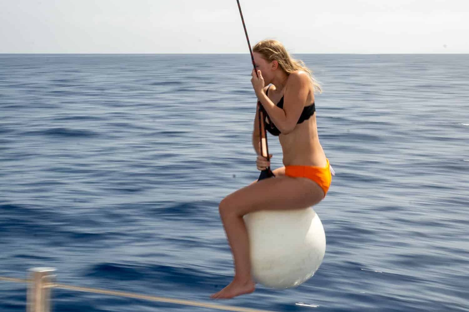 Eine junge Frau im orangefarbenen Bikini sitzt auf einer an einer Stange befestigten weißen Boje über dem tiefblauen Meer und konzentriert sich während ihres Segelurlaubs auf das Angeln.