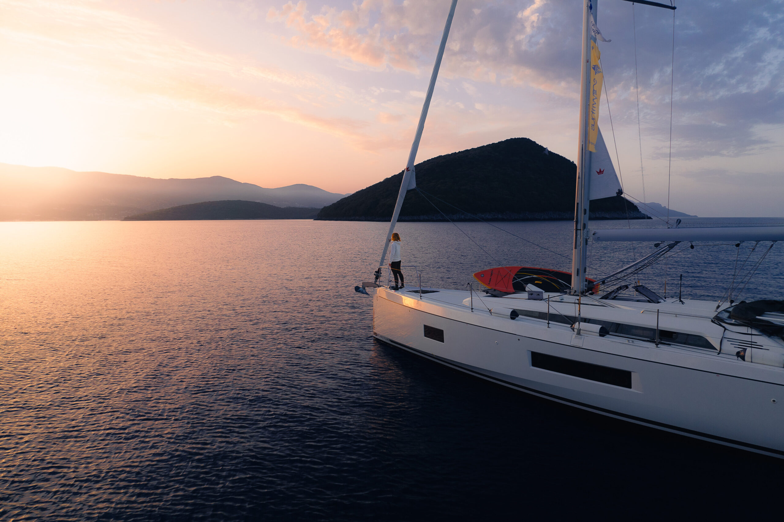 Eine Person steht bei Sonnenuntergang am Bug eines Segelboots auf ruhigem, offenem Wasser. Der Himmel ist eine Mischung aus warmen Farben, am Horizont zeichnet sich eine entfernte Insel ab. Die Szene ruft ein Gefühl von Ruhe und Abenteuer hervor und verkörpert die Essenz eines perfekten Segelurlaubs.