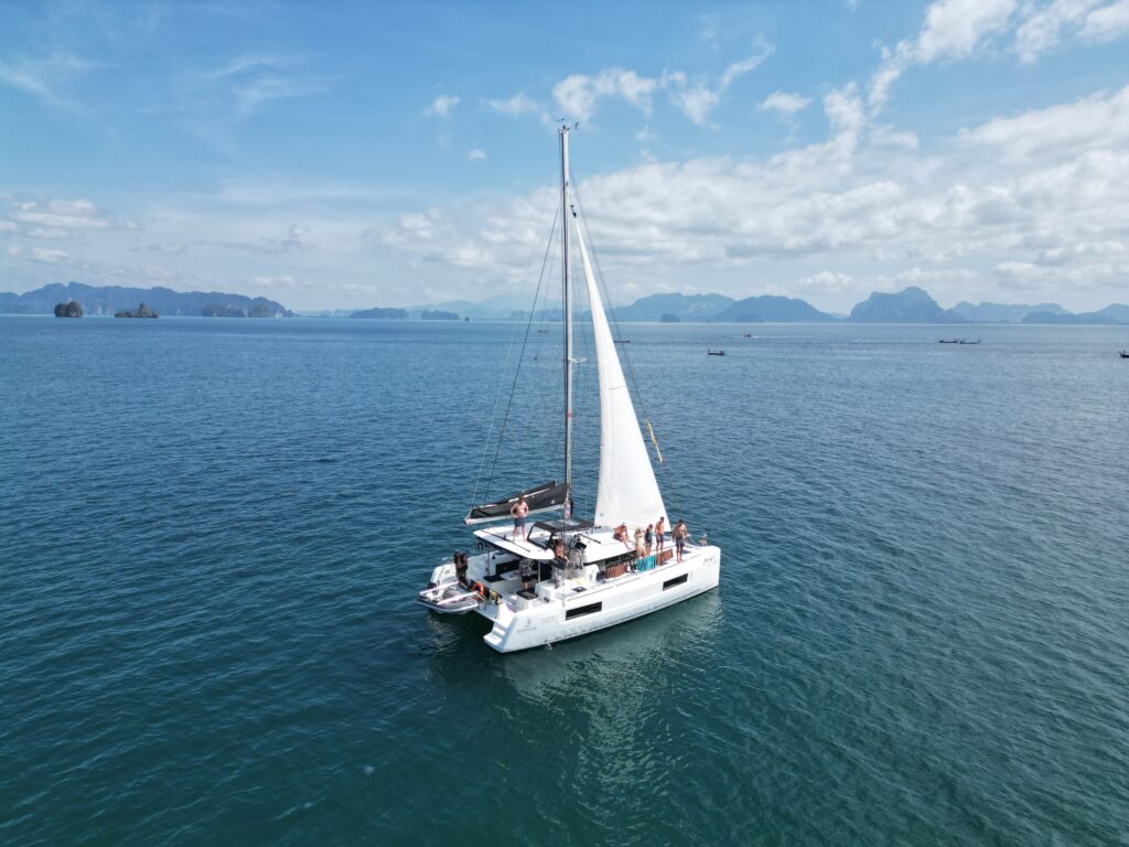 Ein weißes Segelboot mit einer Gruppe von Menschen an Bord kreuzt in den ruhigen blauen Gewässern vor Phuket, Thailand, mit einer Kulisse aus fernen Inseln unter einem teilweise bewölkten Himmel. Das Segel ist vollständig ausgefahren und die Szene zeigt ein ruhiges und malerisches Segeln. Segeln und Windrichtungen: So navigierst du wie ein Profi