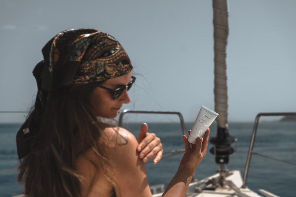 Eine Frau mit buntem Kopftuch und Sonnenbrille sitzt während ihrer Segelreise in Griechenland auf einem Boot und cremt ihre Schulter mit Sonnencreme ein. In der anderen Hand hält sie eine Tube Sonnencreme. Im Hintergrund ist das Meer zu sehen.