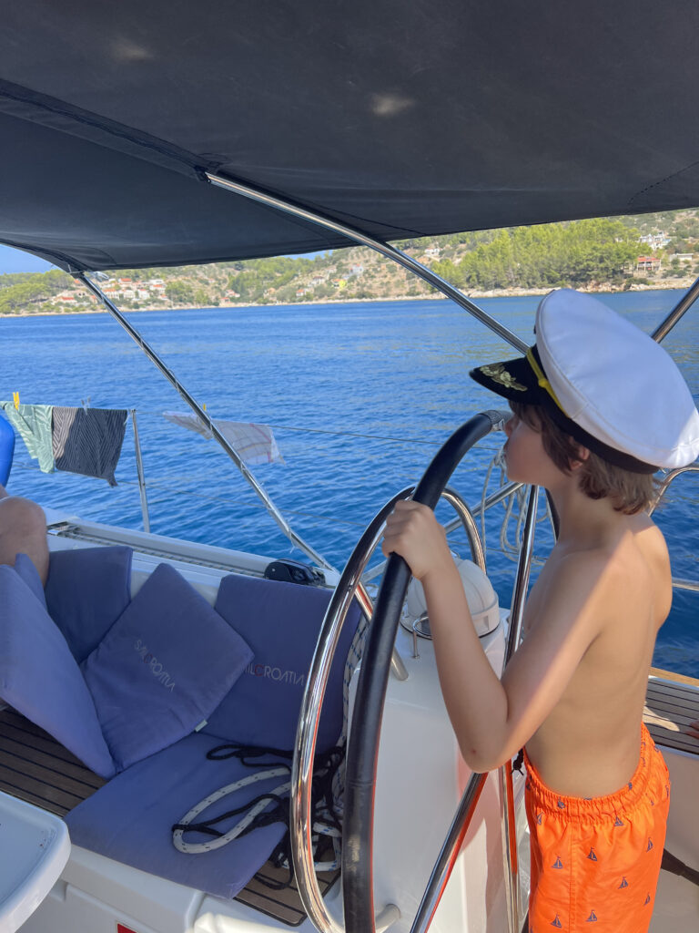 Ein kleiner Junge mit Kapitänsmütze und orangefarbener Badehose steht am Steuer eines Bootes und tut so, als würde er steuern. Das Boot segelt auf klarem, blauem Wasser mit einer hügeligen Küste im Hintergrund und verkörpert den Geist des familienfreundlichen Segeltörns im Mittelmeer. Handtücher und Kleidung trocknen an Bord auf einer Wäscheleine.