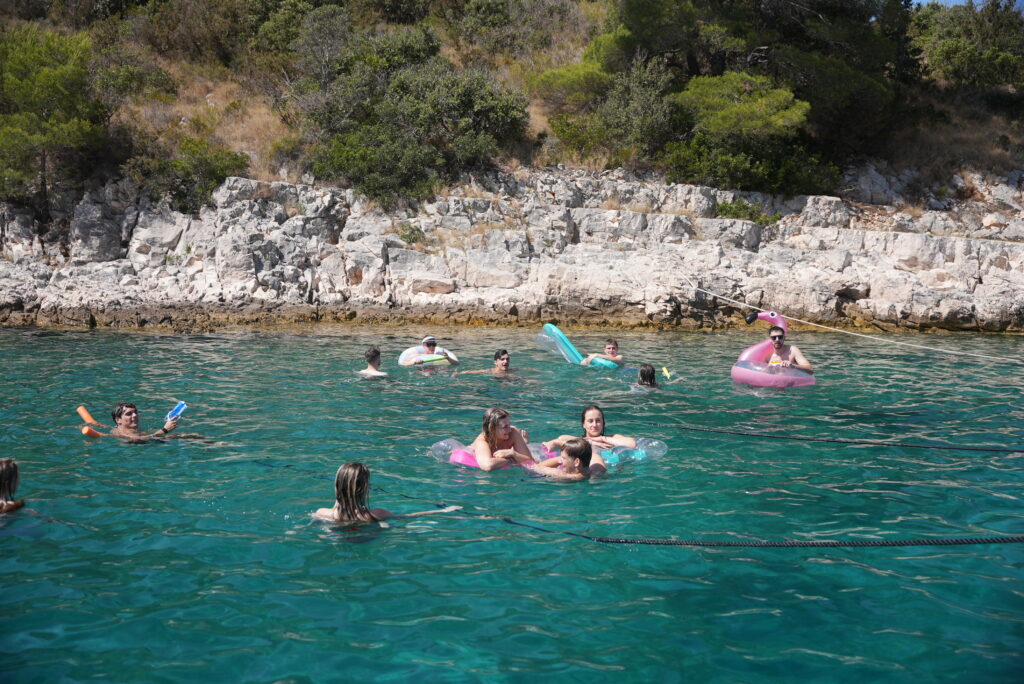 Eine Gruppe von Menschen genießt einen sonnigen Tag im klaren blauen Wasser nahe einer felsigen Küste. Einige schwimmen, während andere bunte Schwimmflügel und Poolnudeln benutzen. Bäume und Sträucher sind auf dem felsigen Hintergrund zu sehen, was an Mitsegeln in Kroatien erinnert. Die Szene ist lebhaft und entspannt. Rundreise plus Badeurlaub: Die perfekte Kombinationsreise