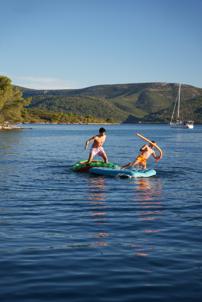 Zwei Kinder spielen auf aufblasbaren Schwimmmatten in einem malerischen See mit ruhigem, klarem blauem Wasser. Ein Kind hält eine Schwimmnudel und scheint spielerisch mit dem anderen zu kämpfen. Im Hintergrund sind bewaldete Hügel und ein Segelboot zu sehen, perfekt für Abenteuer bei Mitsegeln Kroatien, unter einem klaren Himmel.