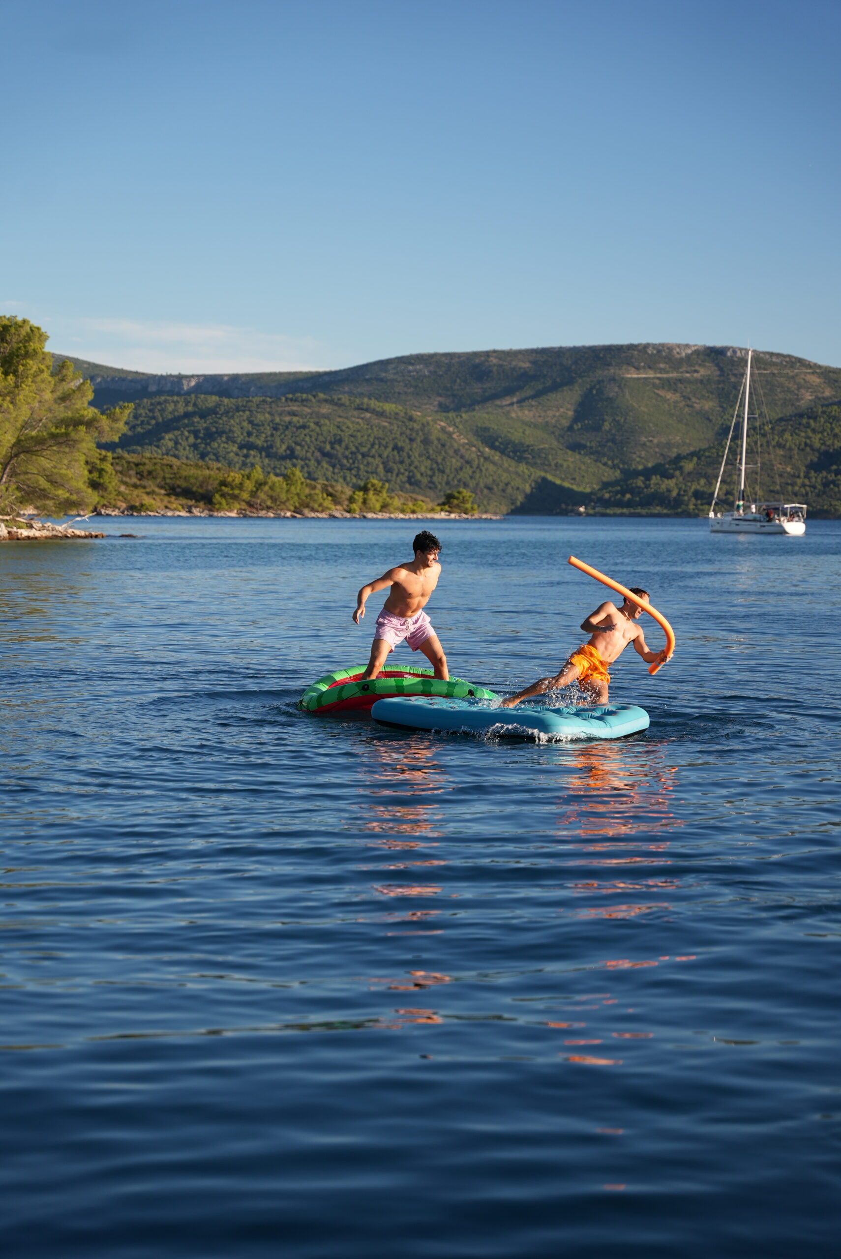 Zwei Kinder spielen auf aufblasbaren Schwimmmatten in einem malerischen See mit ruhigem, klarem blauem Wasser. Ein Kind hält eine Schwimmnudel und scheint spielerisch mit dem anderen zu kämpfen. Im Hintergrund sind bewaldete Hügel und ein Segelboot zu sehen, perfekt für Abenteuer bei Mitsegeln Kroatien, unter einem klaren Himmel.