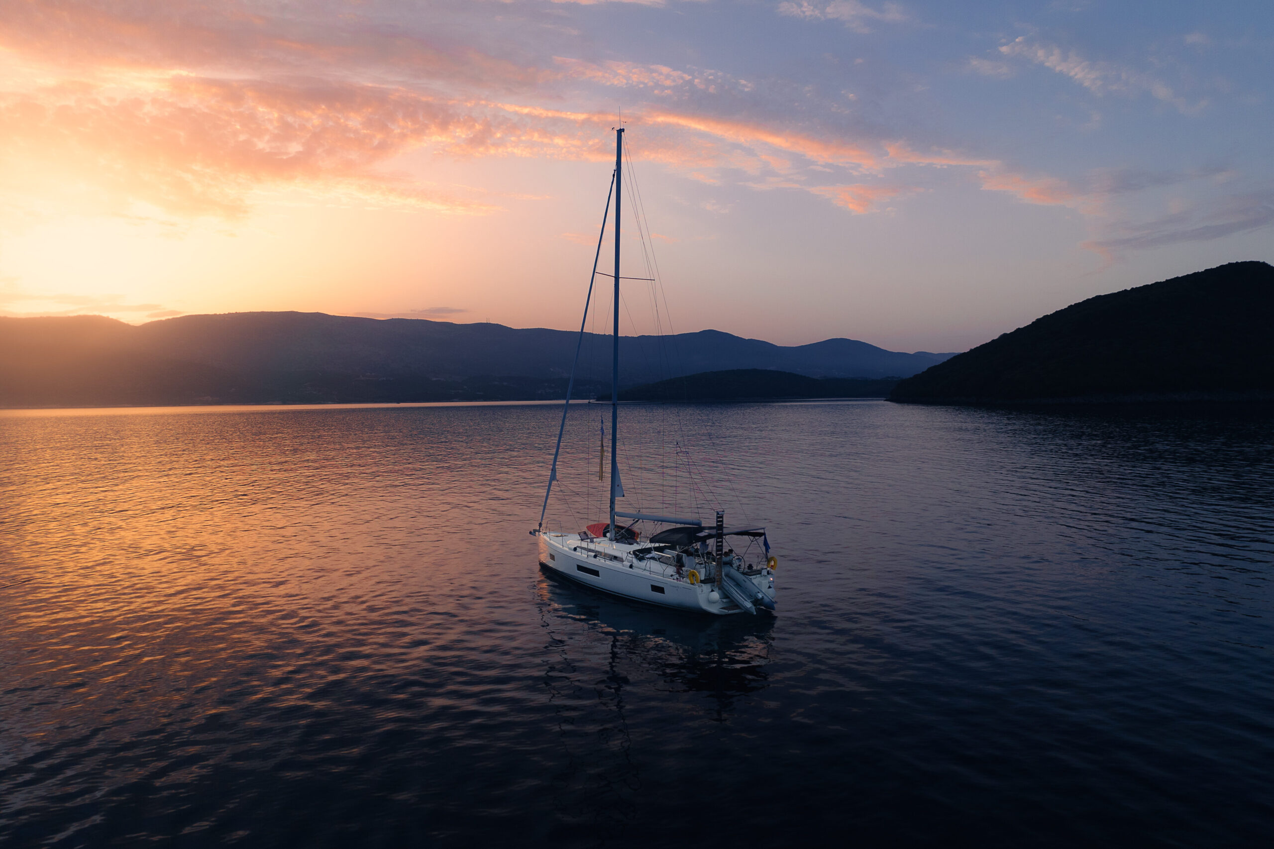 Ein Segelboot schwimmt friedlich auf ruhigem Wasser während eines leuchtenden Sonnenuntergangs und träumt vielleicht von einer Atlantiküberquerung. Der Himmel ist in Orange-, Rosa- und Lilatönen gefärbt. Im Hintergrund zeichnen sich Berge und Hügel ab, was zu der ruhigen und malerischen Szene beiträgt.