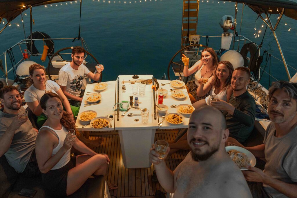 Eine Gruppe von zehn Personen sitzt auf einer Segeltour um einen Tisch und genießt gemeinsam eine Mahlzeit. Auf dem Tisch stehen Teller mit Pasta und Getränken. Die Gruppe scheint sich zu unterhalten und zu lächeln, während festliche Lichterketten eine warme Atmosphäre schaffen, während die Sonne über dem Wasser untergeht. Typisches Bordleben, das dich auf einem Segeltörn erwartet.