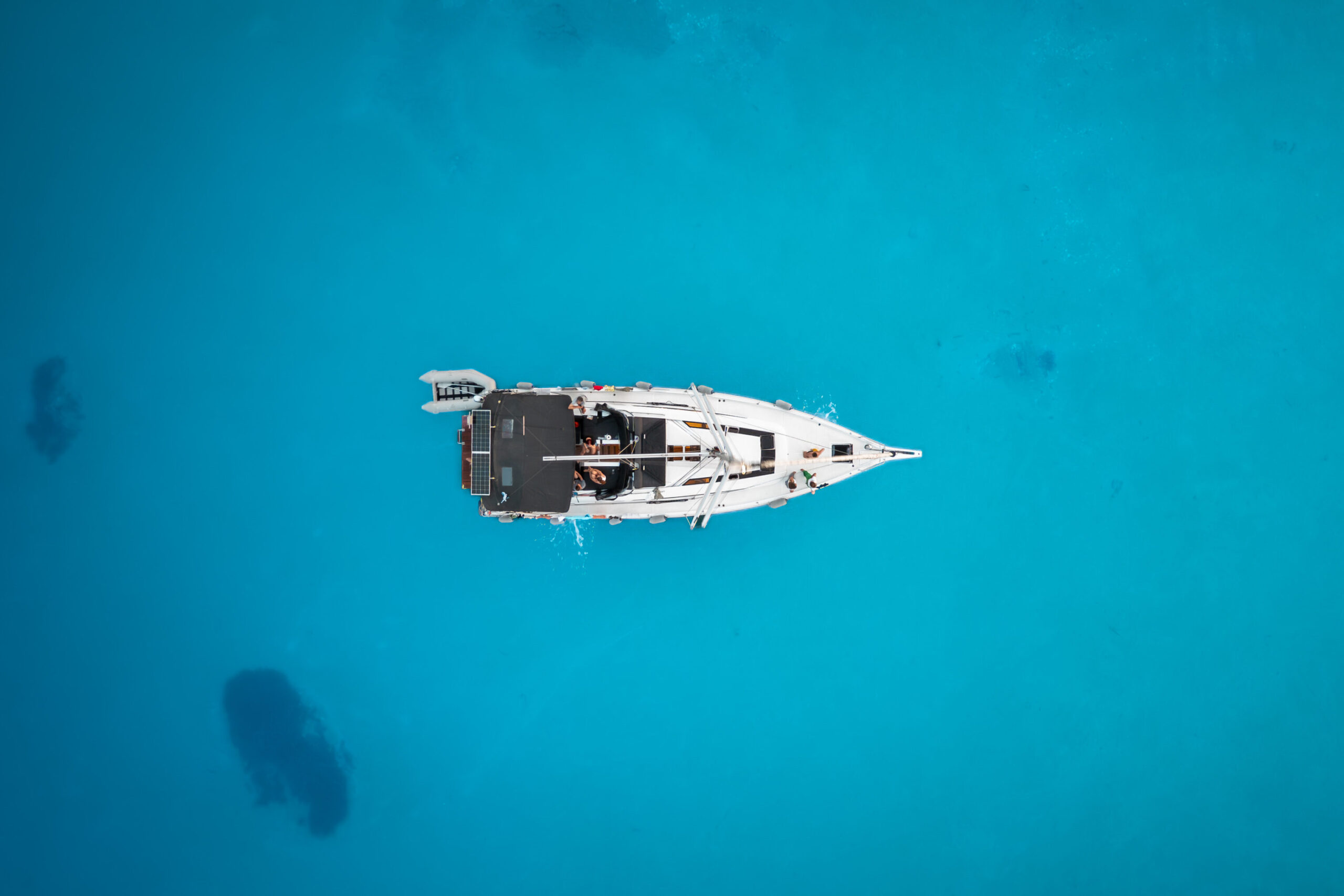 Luftaufnahme eines einzelnen weißen Segelboots, das in klarem, leuchtend blauem Wasser schwimmt. Das Boot befindet sich in der Bildmitte, umgeben von der unendlichen Weite des Meeres, und zeigt seine Ruhe und Abgeschiedenheit – eine heitere Segeltour auf ruhigem Wasser.