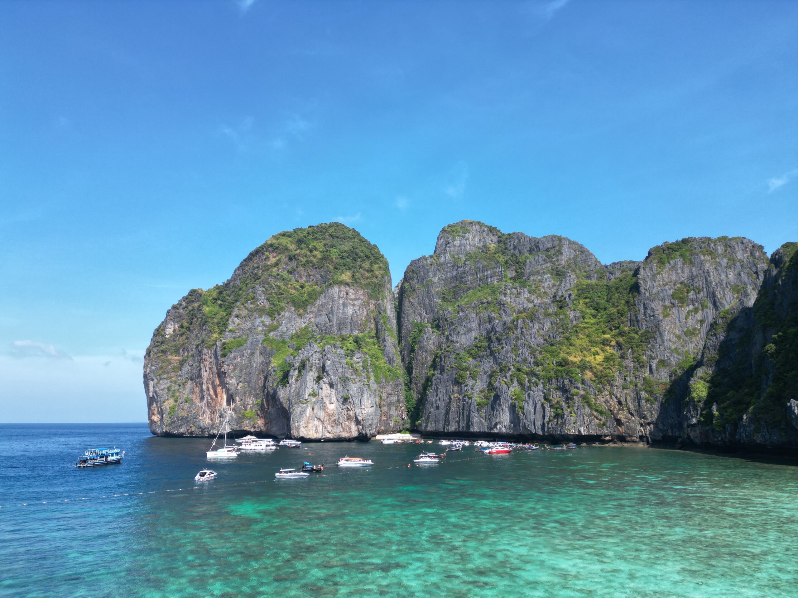 Klares türkisfarbenes Wasser mit mehreren Booten auf einer Segelreise nach Griechenland in der Nähe einer dramatischen Küstenlinie mit üppigen, grünen Klippen, die aus dem Meer ragen. Der Himmel ist strahlend blau mit ein paar vereinzelten Wolken. Die Szene fängt eine tropische, idyllische Küstenlandschaft ein. Thailand Segeltörn als Geheimtipp
