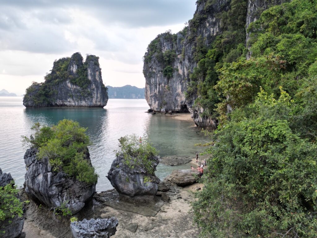 Eine ruhige Küstenszene mit hoch aufragenden Kalksteinklippen, die von üppigem Grün bedeckt sind. Das ruhige, türkisfarbene Wasser plätschert sanft gegen Felsvorsprünge und einen kleinen Sandstrand. Zwei Menschen auf einer Segelreise nach Griechenland sind in der Nähe des Ufers zu sehen, versunken in die natürliche Schönheit der Landschaft. Fernreise planen Traum