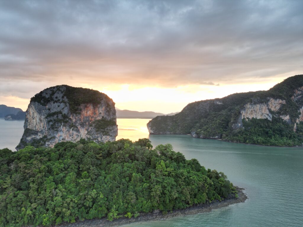 Luftaufnahme von üppig grünen Inseln, umgeben von ruhigem grünem Wasser bei Sonnenuntergang. Der Himmel ist teilweise bewölkt und die Sonne wirft einen goldenen Farbton über die Landschaft. Perfekt für eine Segelreise nach Griechenland. Die Szene umfasst eine große, bewaldete Felsformation im Vordergrund und weitere Inseln im Hintergrund.