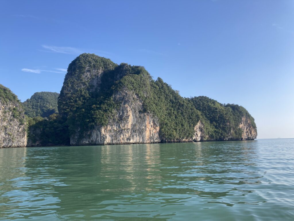Ein heiterer Blick auf ein ruhiges, grünlich-blaues Meer mit großen, üppigen, grün bewachsenen Kalksteinklippen unter einem klaren blauen Himmel, der an eine Segelreise nach Griechenland erinnert. Die Sonne beleuchtet Teile der Klippen, hebt ihre Texturen hervor und erzeugt Spiegelungen im Wasser.