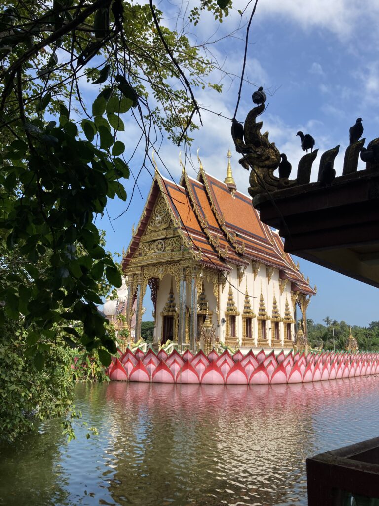 Ein thailändischer Tempel mit reich verzierten goldenen Verzierungen und einem roten Dach steht auf einer von Wasser umgebenen Plattform und erinnert an die heiteren Ausblicke, die man auf einer Segelreise nach Griechenland erleben kann. Der Sockel des Tempels ist mit rosa Lotusblüten geschmückt, während üppiges Grün und eine kunstvolle Struktur mit Vogelskulpturen den Vordergrund unter einem klaren blauen Himmel zieren.