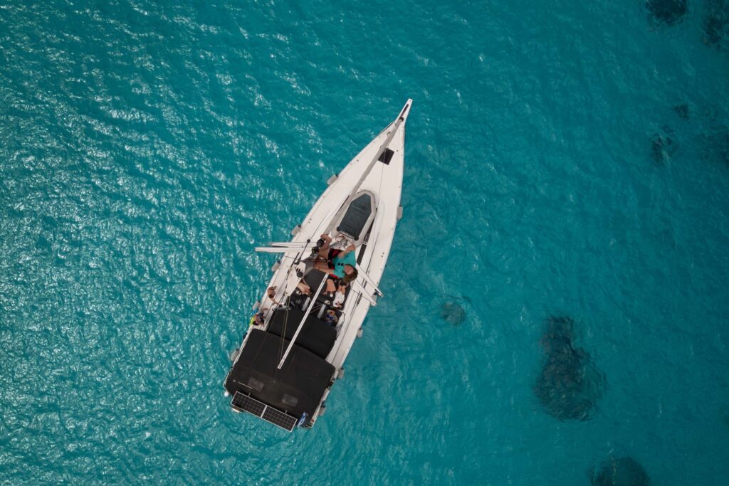 Luftaufnahme eines Segelboots mit einigen Personen an Deck, die während ihrer Segelreise Griechenland in klarem türkisfarbenem Wasser treiben. Der weiße Rumpf des Bootes kontrastiert mit dem leuchtend blauen Meer und schafft eine ruhige und malerische Szene.